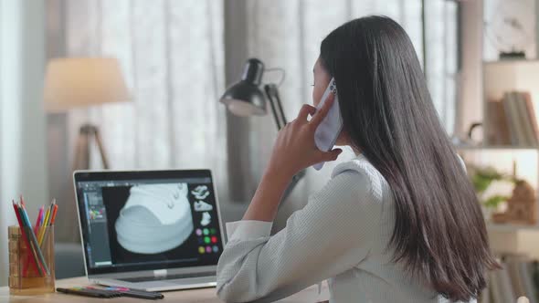 Asian Female Footwear Designer Talking On Smartphone While Designing Shoe On A Laptop At Home