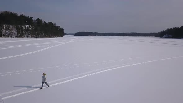 Rotating aerial shot around blonde girl wandering on a vast open area covered in snow.