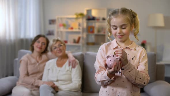 Schoolgirl Putting Coin in Piggybank, Proud Mother and Granny Behind, Savings