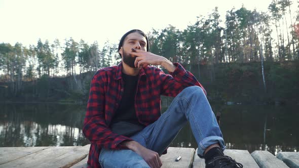 Bearded Yung Man Sit Near Amazing Lake and Smoke Cigar