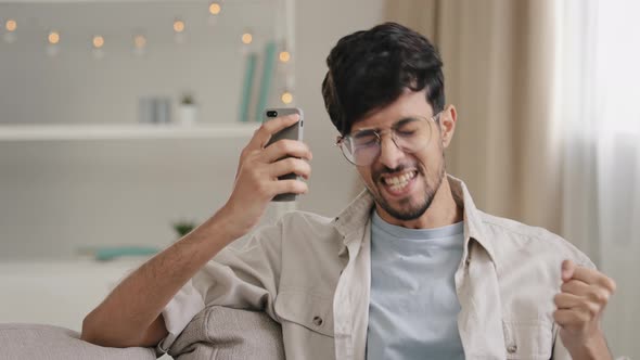 Young 30s Arab Bearded Man Millennial Guy in Glasses Sits at Home on Couch Looking at Mobile Phone