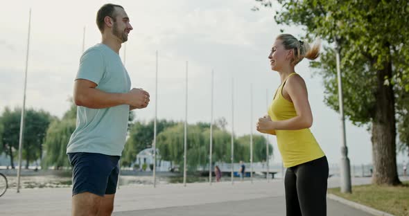 Happy Couple Squatting on Embankment. Side View of Cheerful Man and Woman in Sportswear Smiling and