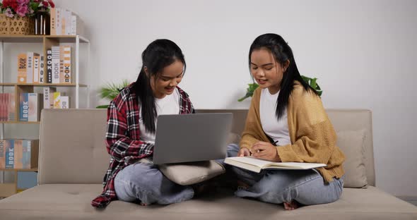 Twin girls reading a book and looking laptop while sitting on couch