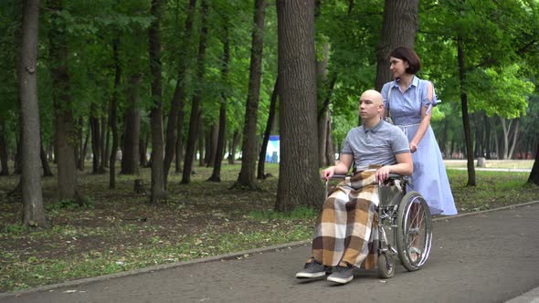 A Woman Is Driving a Young Man with Oncology in a Wheelchair Through the Park. The Man Is Bald Due