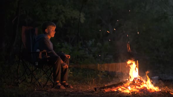 Child boy sitting near capming fire with bright sparks at night.