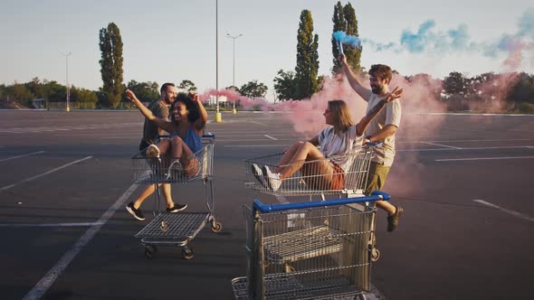 Young Men are Laughing Holding Smoke Bombs and Riding Shopping Carts with Their Girlfriends at