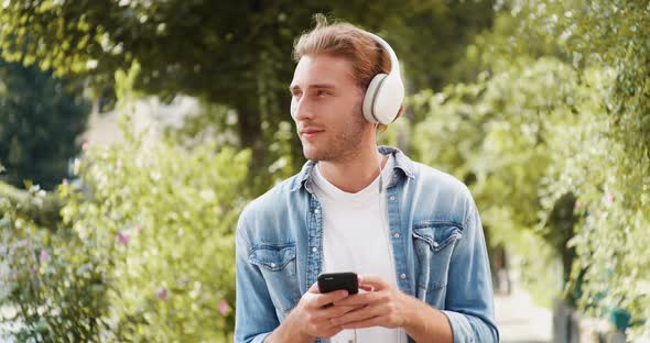 Cute Caucasian Man is walking on Sunny Day in Headphones, surfing the Internet.