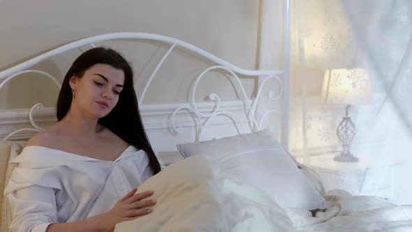 a Brunette in a White Shirt and with Long Hair is Sitting in a Bed in a Light Room