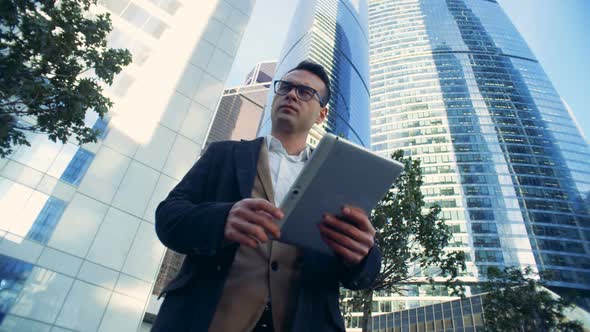 Male Businessman, Manager Is Standing Near Skyscrapers with a Tablet.