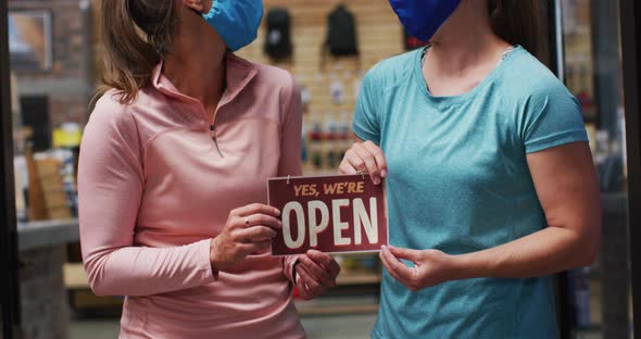 Portrait of caucasian female shopkeepers in face masks with open sign in window of sports shop