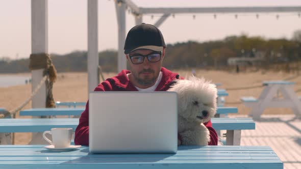 Man with a Dog in a Cafe Works Remotely on a Laptop in a Resort