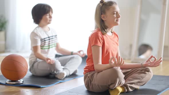 Side View of Concentrated Caucasian Girl Sitting in Lotus Pose with Closed Eyes and Blurred Boy