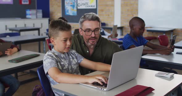 Diverse male teacher helping a schoolboy sitting in classroom using laptop