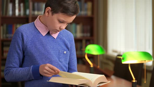 Portrait Shot of the Cute Schoolboy and Standing Near the Bookshelf in the Library and Reading