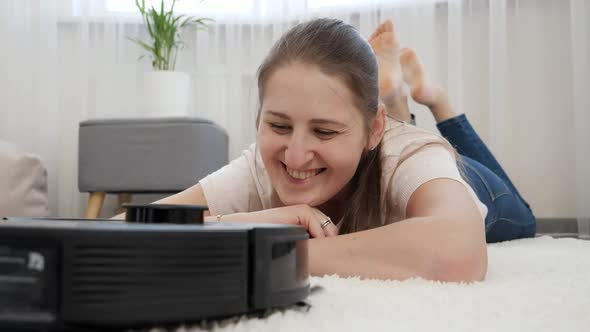 Portrait of Happy Smiling Woman Lying on Carpet and Looking on Working Robot Vacuum Cleaner