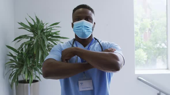 Portrait of african american male doctor wearing scrubs and face mask, crossing arms in hospital