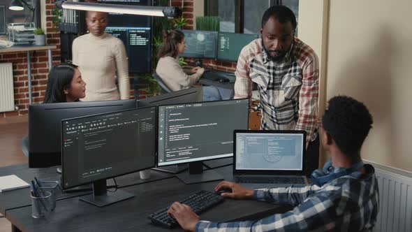 Programer Sitting at Desk with Multiple Screens Running Code Talking with Colleague