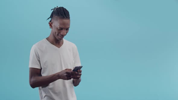 Black man smiling uses phone and emotionally rejoices while standing on a blue studio background.