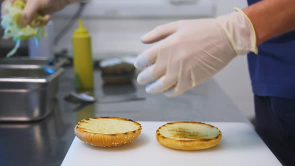 Chef makes a burger, puts a fresh green leaf of lettuce on a bun close up view. 