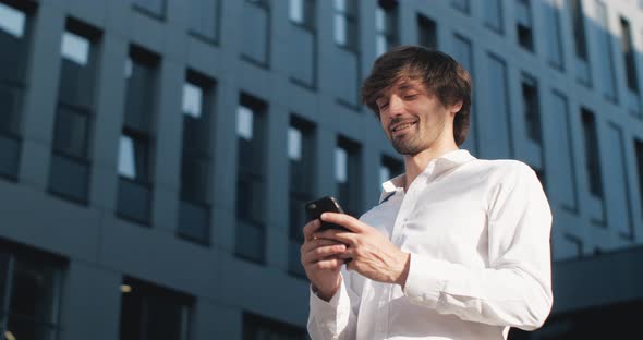 Young successful Businessman is texting Messages on his Smartphone near Office Building.