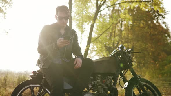 Young Attractive Man in Leather Jacket and Stylish Sunglasses is Standing By His Bike on the Country