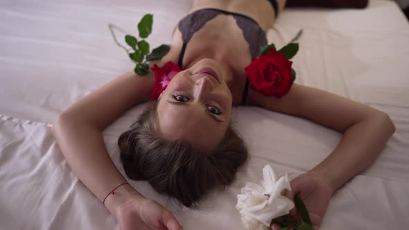 Young Passionate Happy Woman Looking at Camera Lying on Bed with Flowers Around