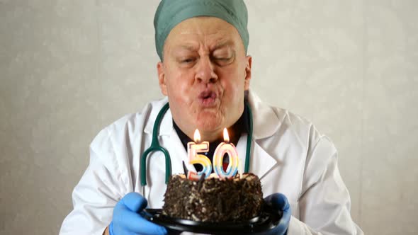 Joyful Mature Adult Doctor in Medical Mask and White Robe, Blows Out Candles on Birthday Cake.