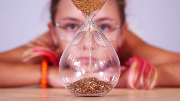 Girl with Pink Hair Looks at Sand Inside Sandglass at Table