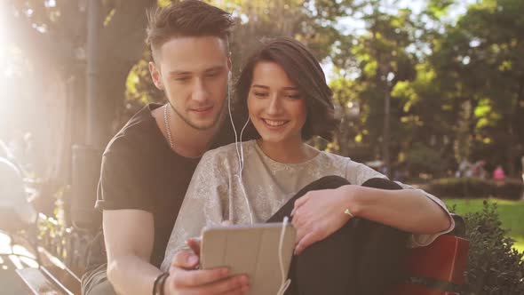 Young Beautiful Couple Smiling Speaking Looking at Tablet Sitting in City Park