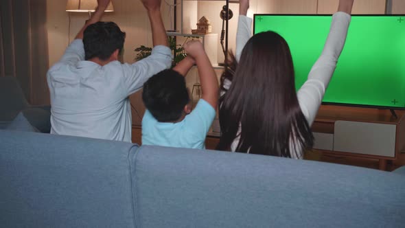 Back View Of Excited Asian Family Watching Tv With Mock Up Green Screen Together Sitting On A Sofa