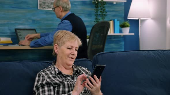 Pleasant Senior Older Lady Resting on Couch Using Applications on Smartphone