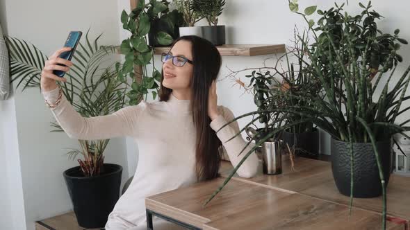 Young Woman with Eyeglasses Making Selfie Stting in Cafe with Many Plants on Background