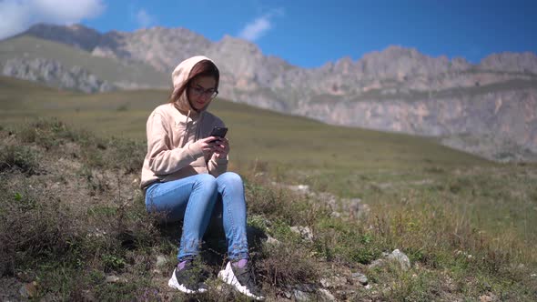 A Young Woman in a Sweatshirt Sits with a Phone in Her Hands Against a Background of Mountains.