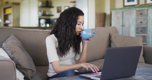 Biracial woman drinking coffee, sitting on sofa and using laptop