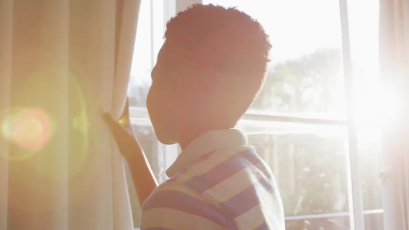 African american boy opening curtain and looking out of sunny window, backlit by sun