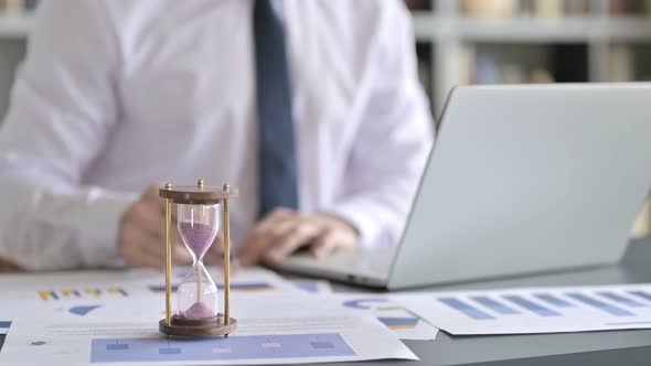 Close Up of Hourglass on Desk and Businessman Working on Laptop