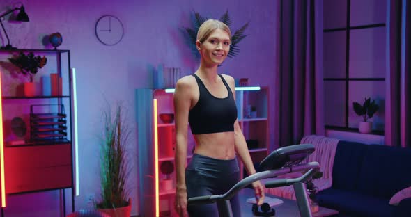 Woman in Sportswear Posing on Camera while Stepping on Treadmill During Evening Home Workout