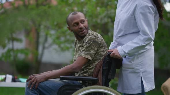 African American Paralyzed Man Greeting Unrecognizable Caucasian Nurse Pushing Wheelchair Leaving