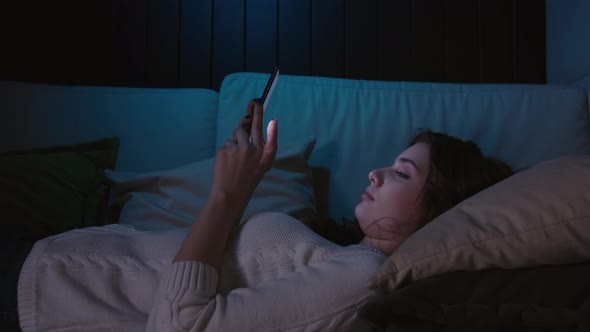Millennial Woman Holding Cellphone Lying On A Couch at Night, Swiping Photos In Online Dating App