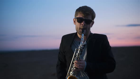 Bearded Man in Black Jacket and Glasses Play Saxophone Outdoors in Mountains on Plain at Dusk at