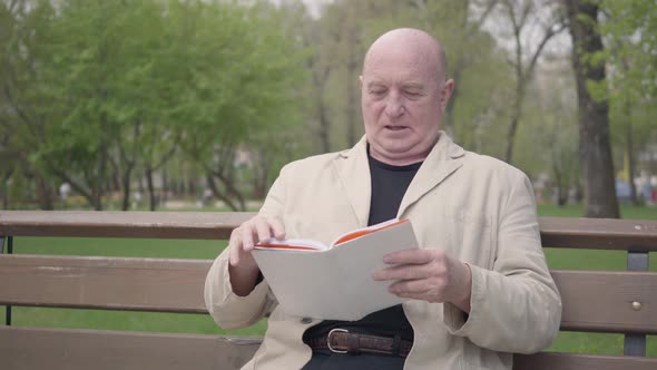 The Bald Mature Man Sitting in the Park on the Bench Reading a Book