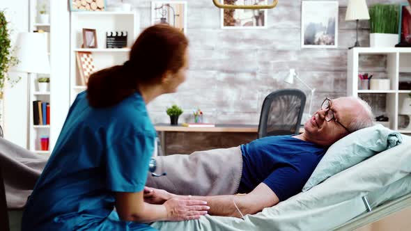 Nurse Taking a Seat Near an Old Sick Man Who Lies in Hospital Bed