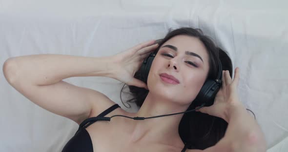 Joyful Girl Lying on Bed Puts on Headphones Sways with Closed Eyes and Looks