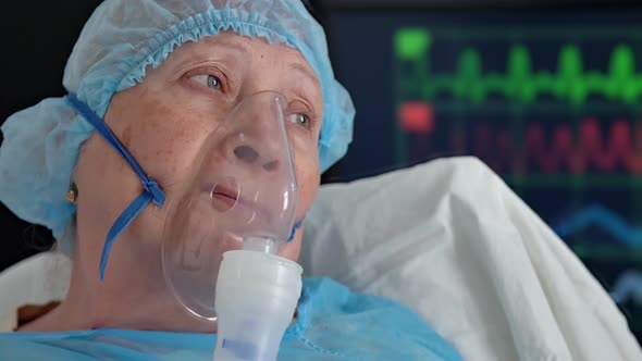 Portrait of Old Woman in Oxygen Mask on Hospital Bed Against Background of Monitor with Vital Signs