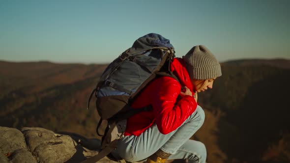 Slow Motion Adventurous Woman Hiker in Red Jacket with Backpack Sits on Cliff Top at Sunset Above