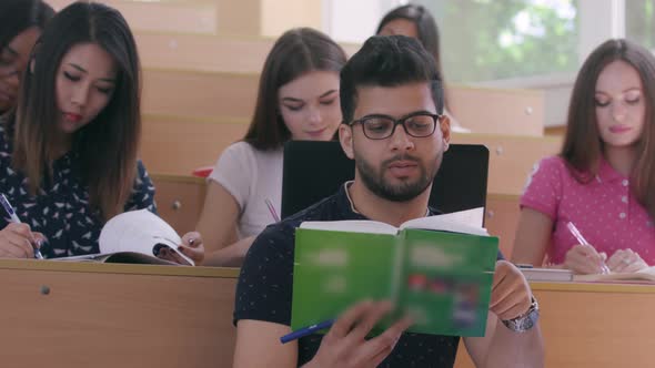 Young Indian Guy Reading Book While Classmates Taking Notes