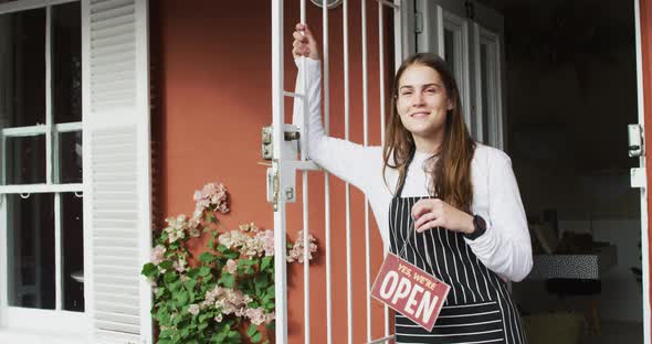 Smiling caucasian waitress standing in door, holding open sign, looking at camera