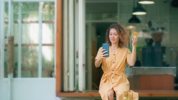 Smiling Curly Woman in Yellow Dress Using Her Phone Happily While Sits in Coffee Shop