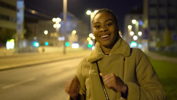 A Young Black Woman Dances and Smiles at the Camera in a Street in an Urban Area at Night