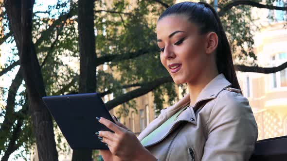 Young Beautiful Woman Sits on the Bench in Park and Works on the Tablet